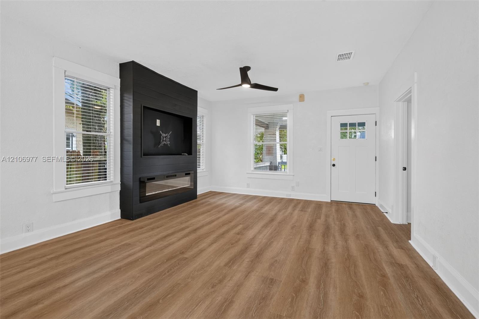 Living area featuring wood-finish flooring, a matte black shiplap accent wall with an integrated electric fireplace, and a ceiling fan with integrated lighting
