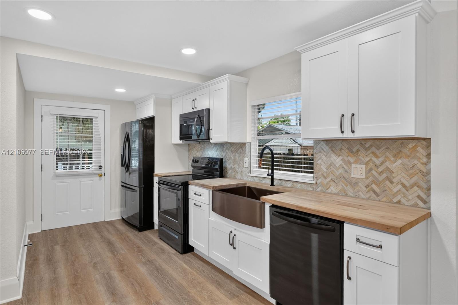 Kitchen featuring white cabinetry, butcher block countertops, a farmhouse-style sink, matte black hardware, and herringbone-patterned tile backsplash