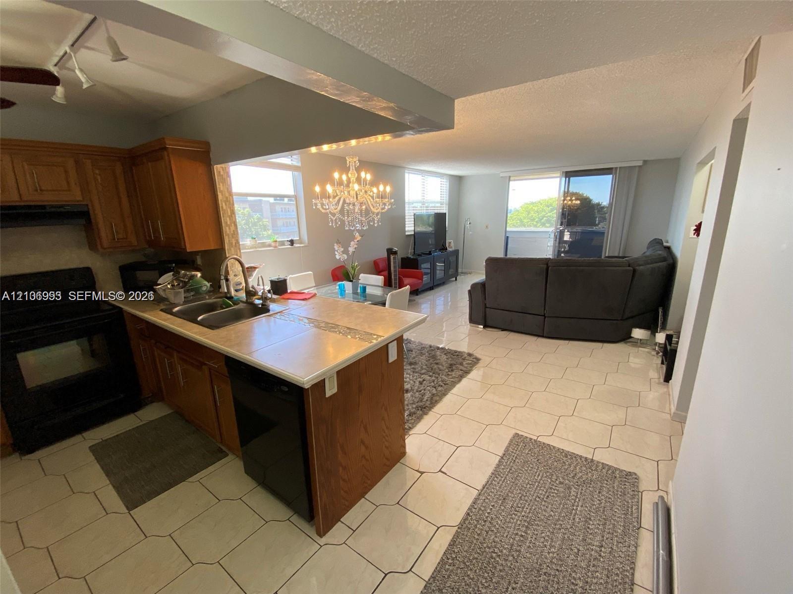 Kitchen featuring wood cabinetry, tile countertops, stainless steel dual-basin sink, and track lighting, complemented by tile flooring and an ornate chandelier