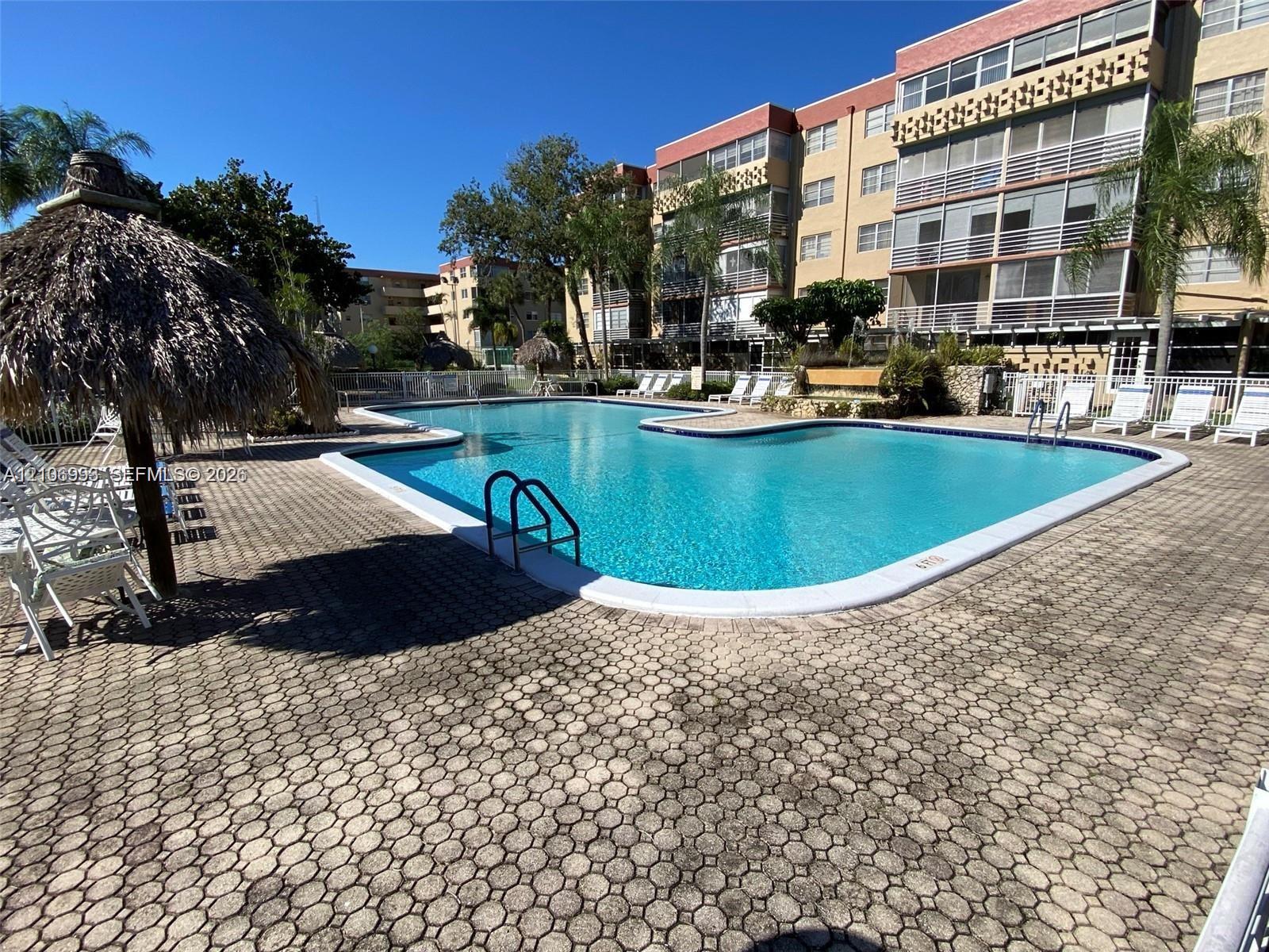Swimming pool with integrated steps, surrounded by a paved patio, thatched-roof cabana, and multi-story building facade