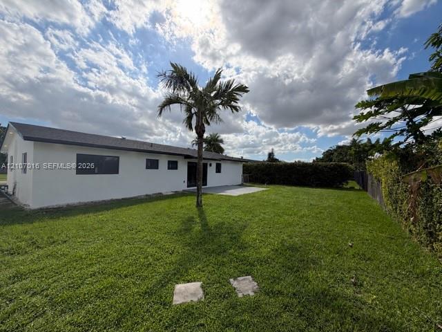 Rear exterior featuring white stucco siding, dark-toned roof shingles, rear concrete patio, expansive grass lawn, and mature palm tree