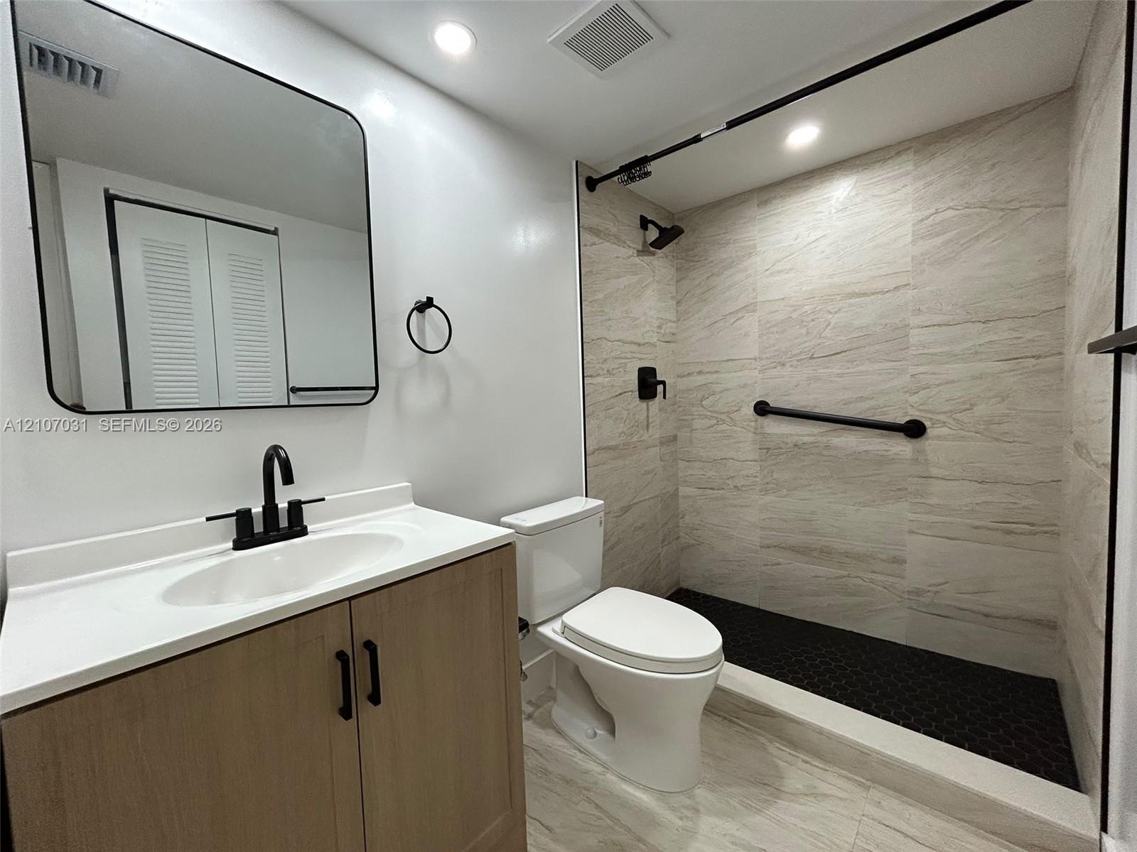 Bathroom featuring a wood-finish vanity with a white countertop, matte black fixtures, a rectangular vanity mirror with rounded corners, and a walk-in shower with stone-patterned wall tile and a black hexagonal tile floor