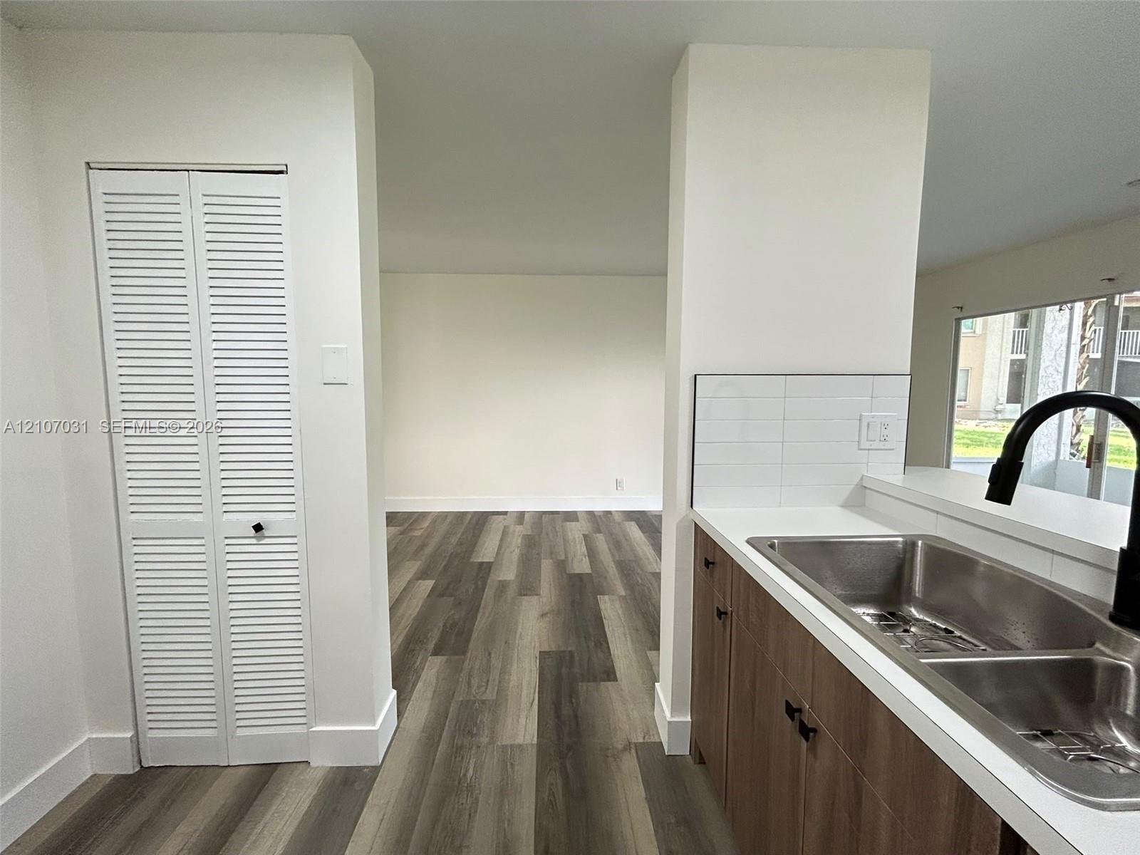 Kitchen featuring wood-finish cabinetry, white countertops, a subway tile backsplash, a stainless steel double-basin sink with a matte black faucet, and wood-finish flooring