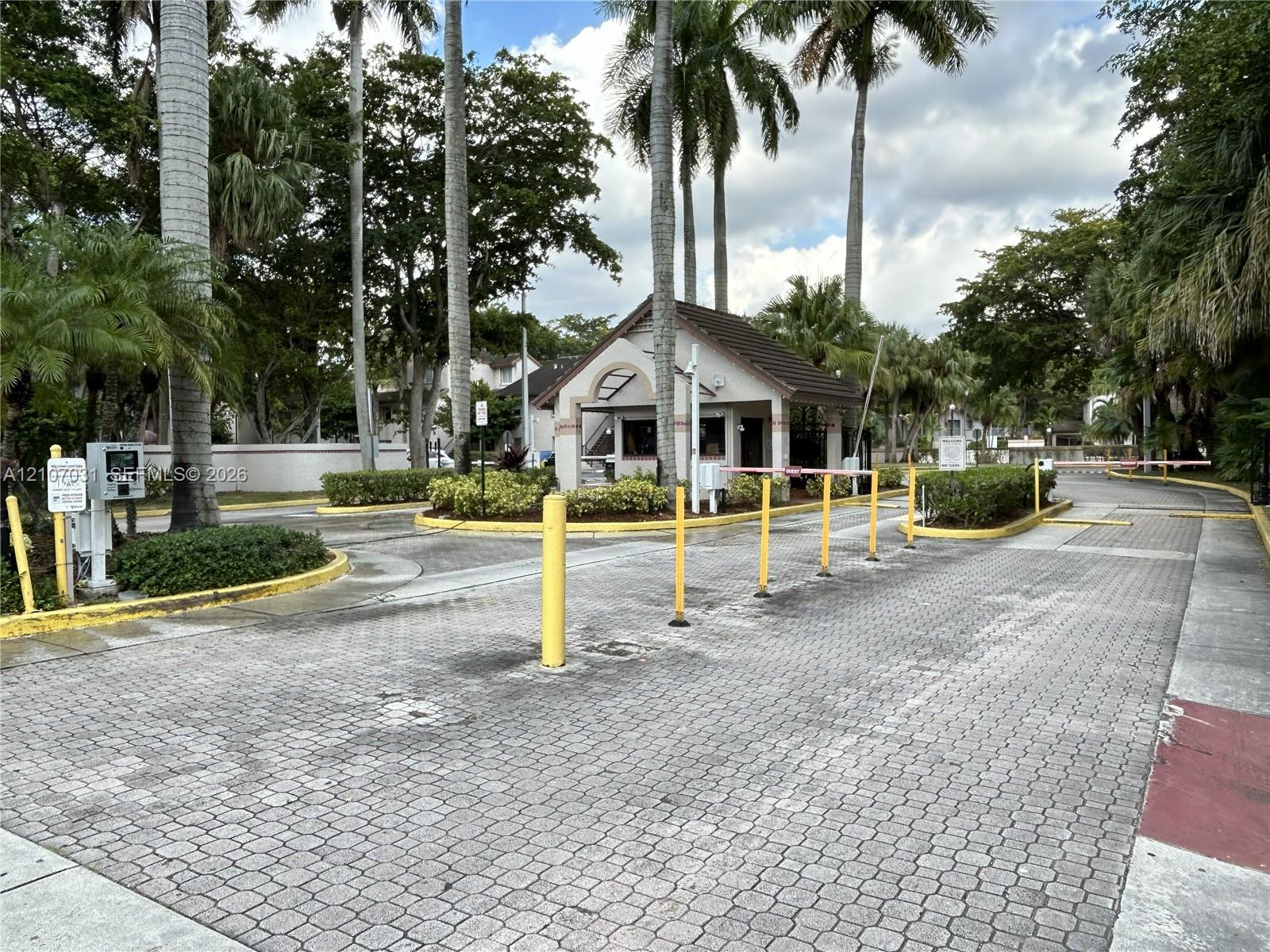 Gated entry featuring a controlled-access security gate, stone paver driveway, guard house, and mature palm landscaping