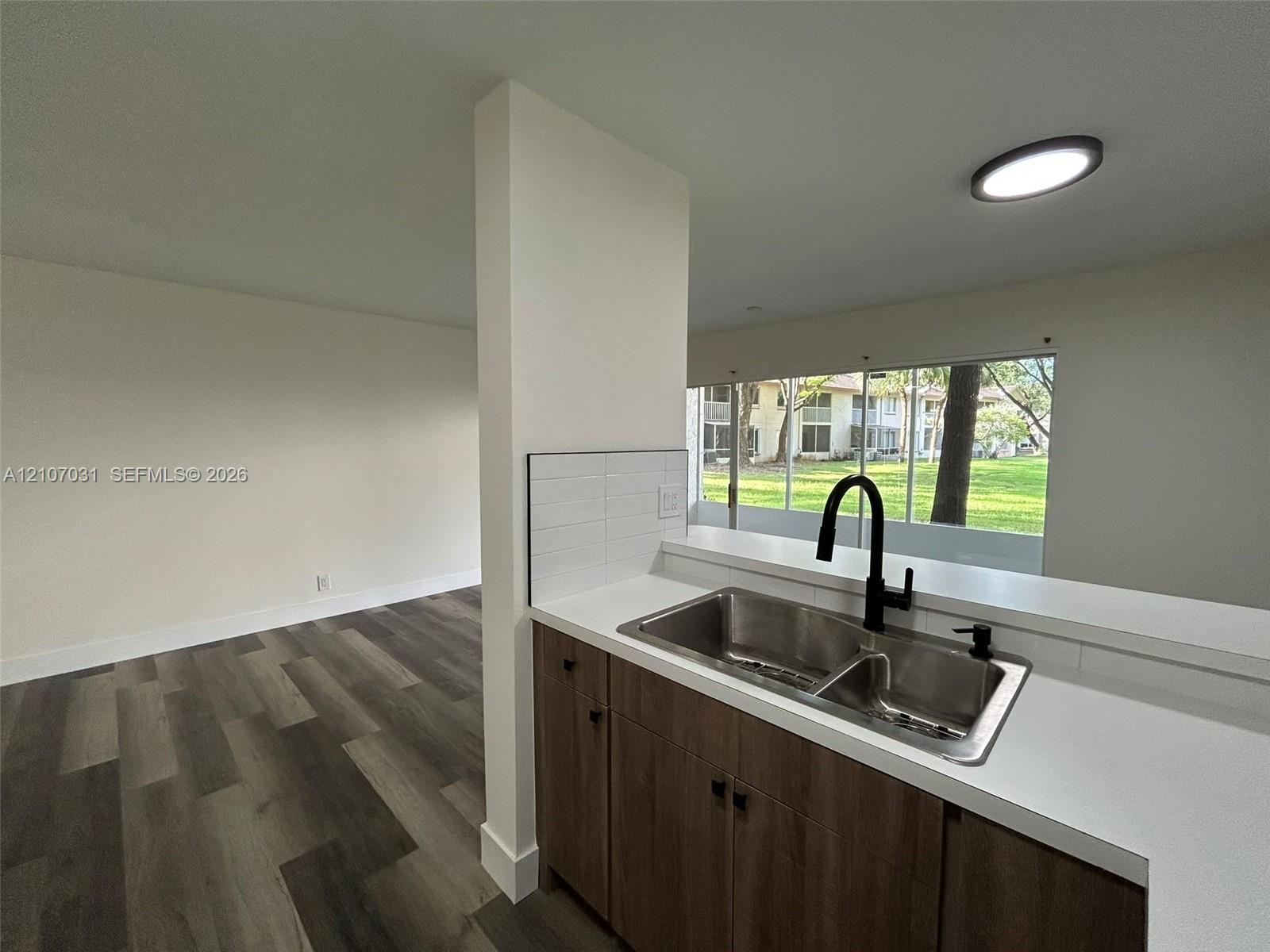 Kitchen featuring wood-finish cabinetry, white solid-surface countertops, a stainless steel dual-basin sink with a matte black high-arch faucet, and wood-finish flooring