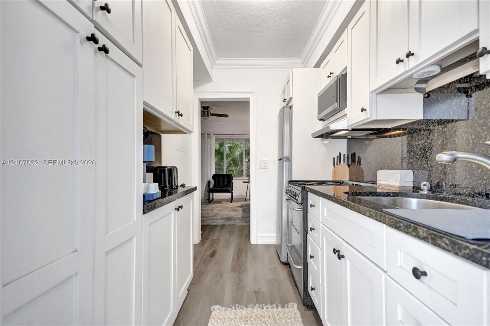 Galley kitchen featuring white shaker-style cabinetry, stone countertops, integrated stainless steel appliances, dark stone backsplash, and wood-finish flooring