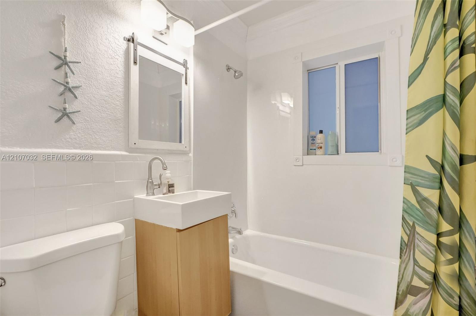 Bathroom featuring a wall-mounted vanity with a rectangular basin, subway tile wainscoting, sliding barn-door style mirror, and a built-in bathtub with shower surround