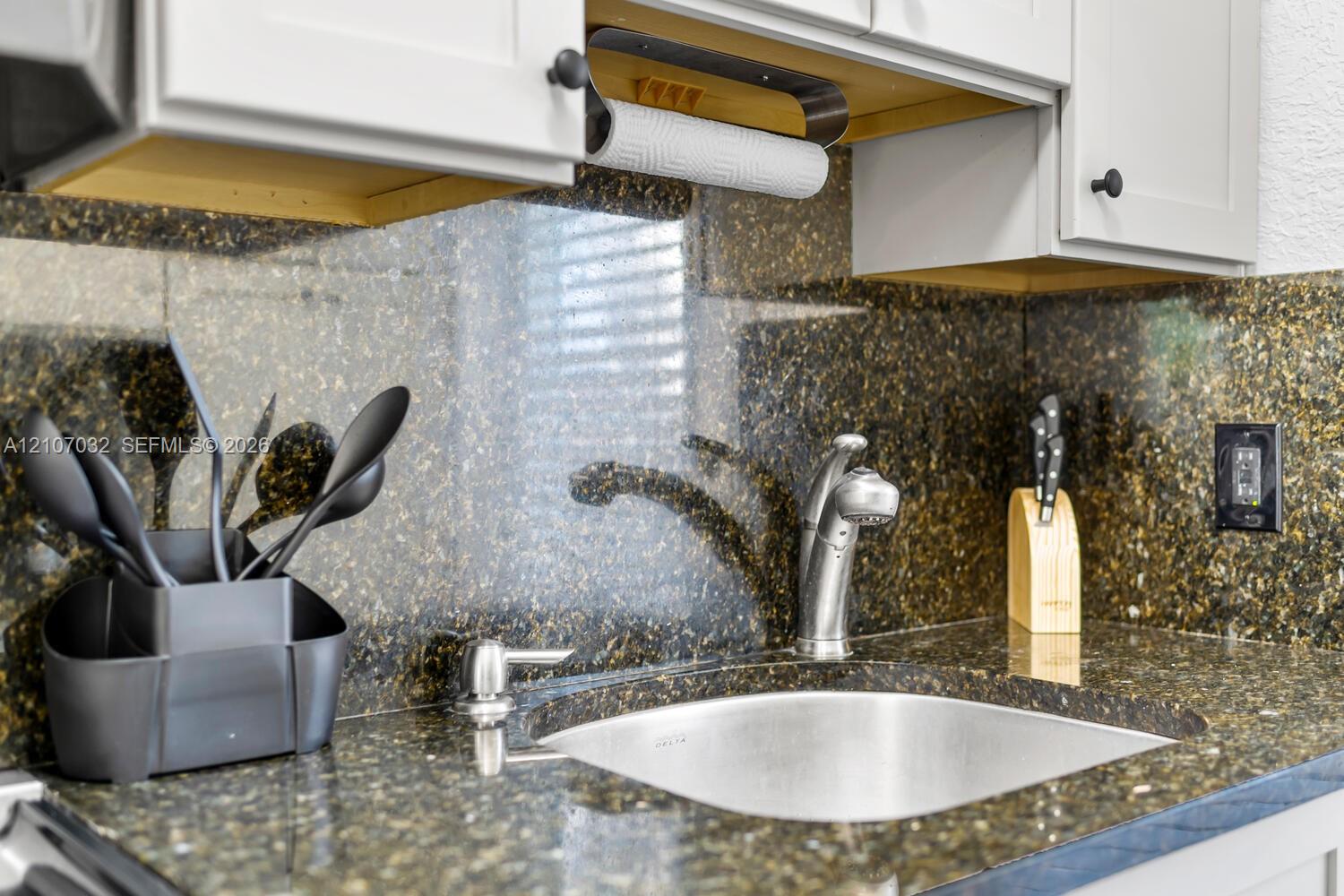 Polished stone countertop and matching full-height backsplash, undermount stainless steel sink, pull-down chrome faucet, and shaker-style cabinetry with matte black hardware