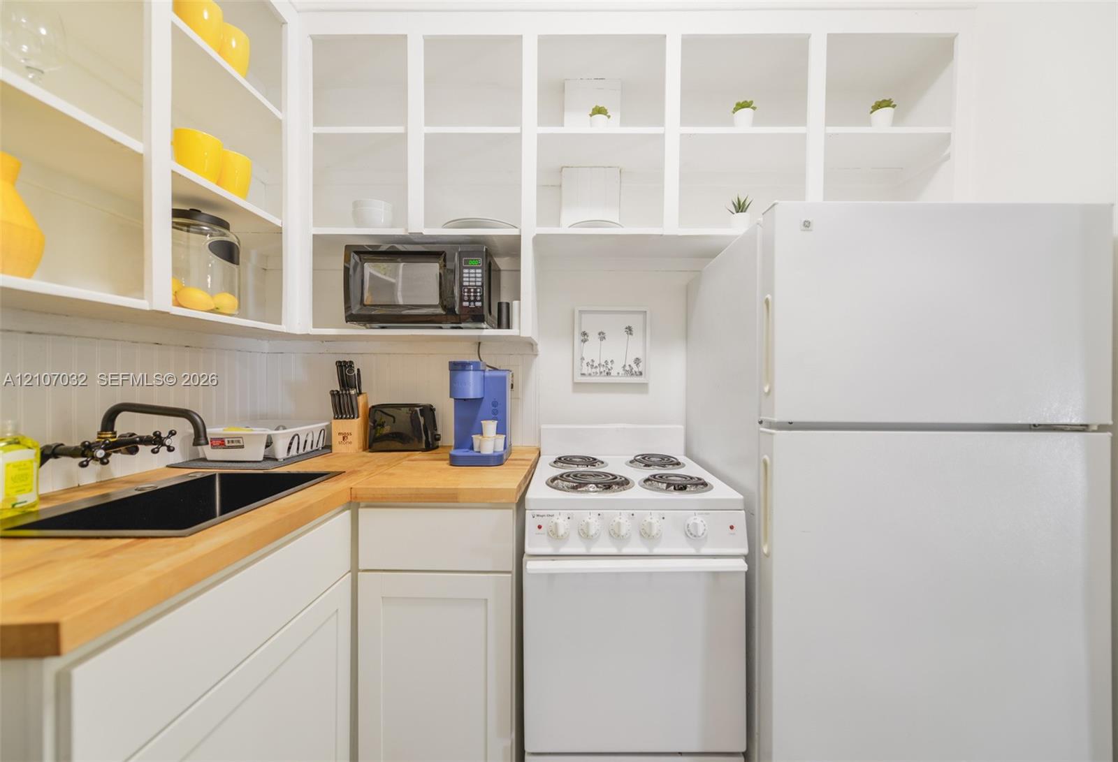 Kitchen featuring white cabinetry with open shelving, wood-finish countertops, an undermount sink with a matte black faucet, and white appliances