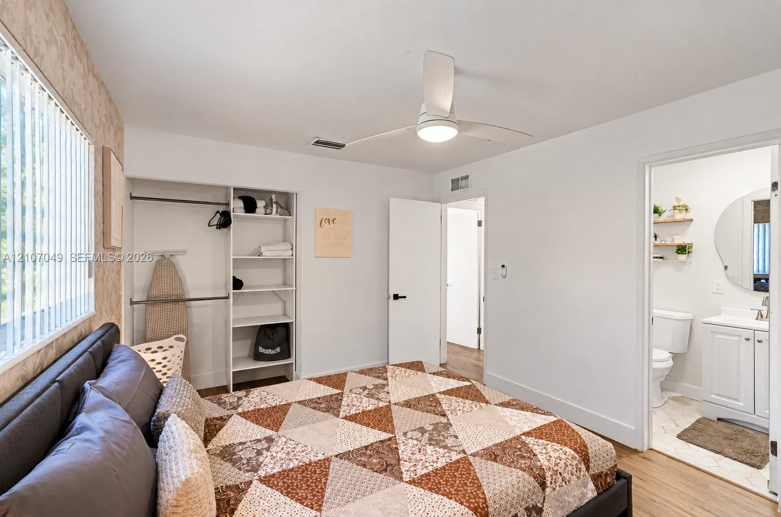 Bedroom featuring wood-finish flooring, a ceiling fan with integrated lighting, a built-in closet system with open shelving, and a bathroom entry with a vanity and wall-mounted circular mirror