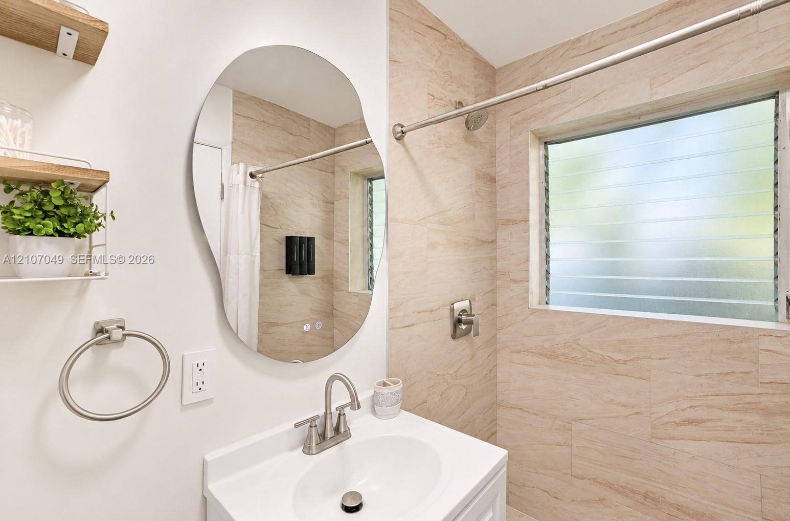 Bathroom featuring a vanity with a white basin, an irregular-shaped wall mirror, louvered glass window, neutral-toned tiled shower surround, and satin nickel fixtures