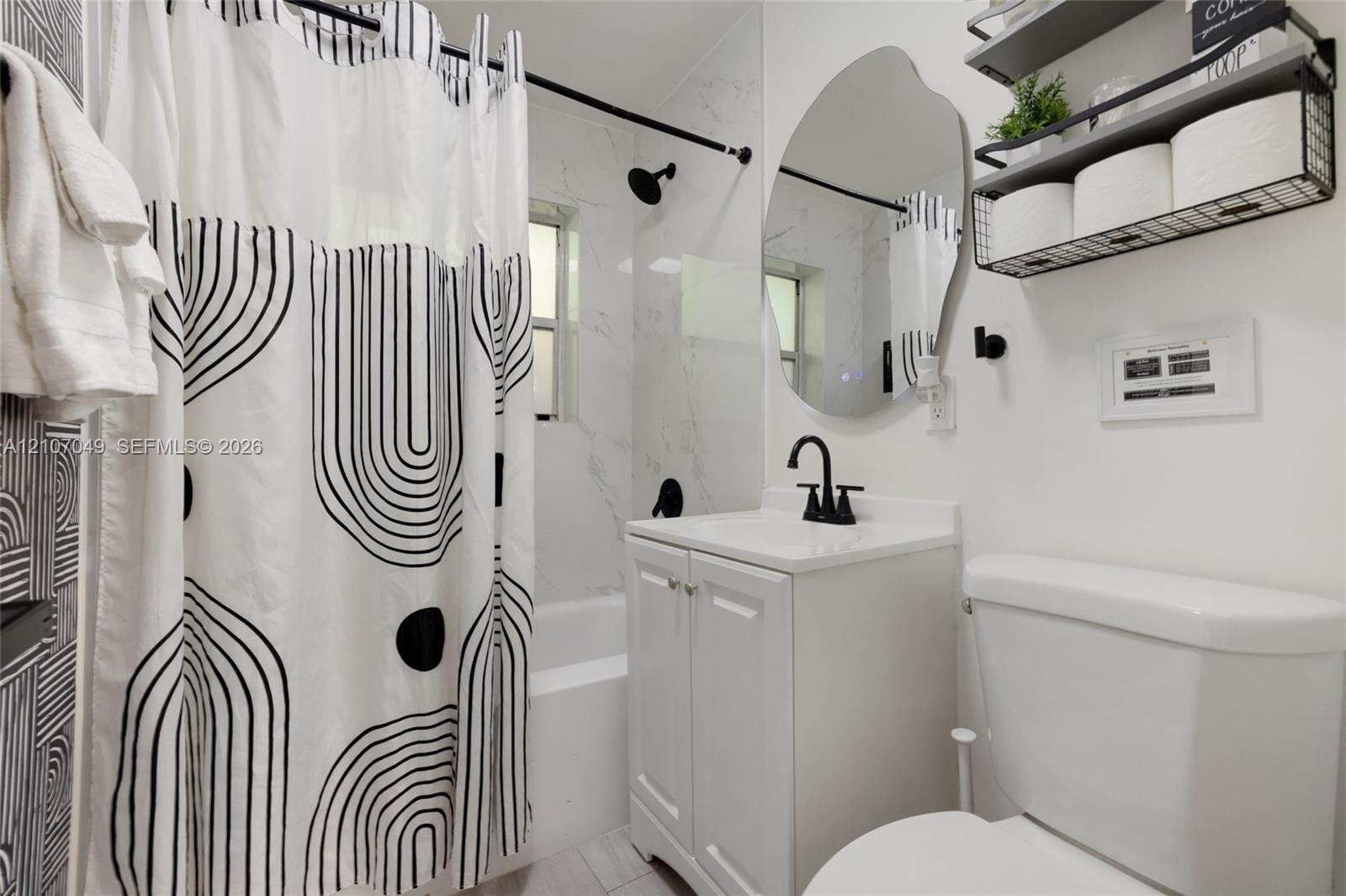 Bathroom featuring a vanity with a white cabinet, stone-finish countertop, and matte black faucet, accompanied by a tub with light-toned wall tile and a matte black shower fixture, complemented by an irregular-shaped wall mirror