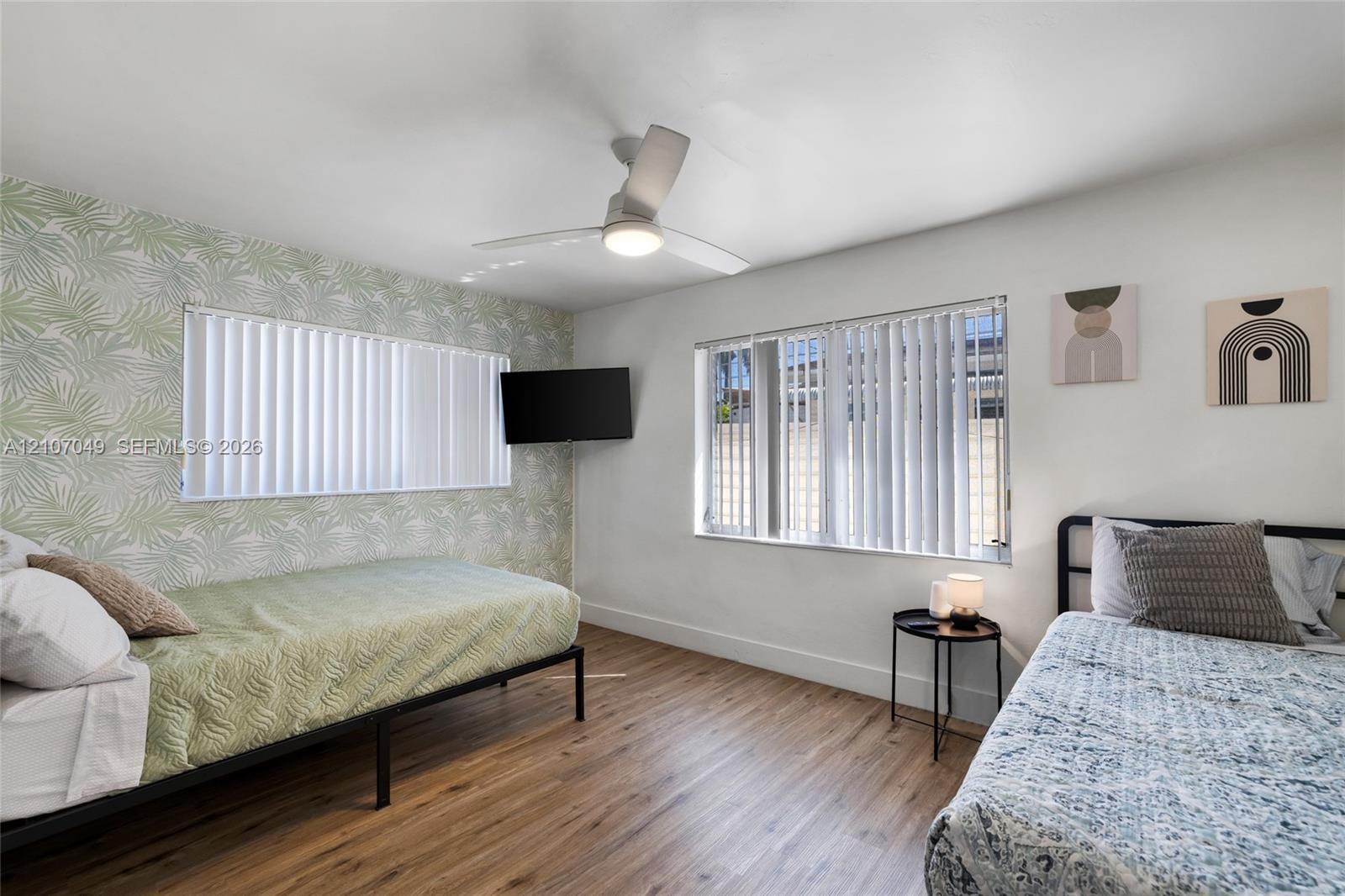 Wood-finish flooring, patterned accent wall, and ceiling-mounted light fixture with integrated fan, featuring dual windows with vertical blinds