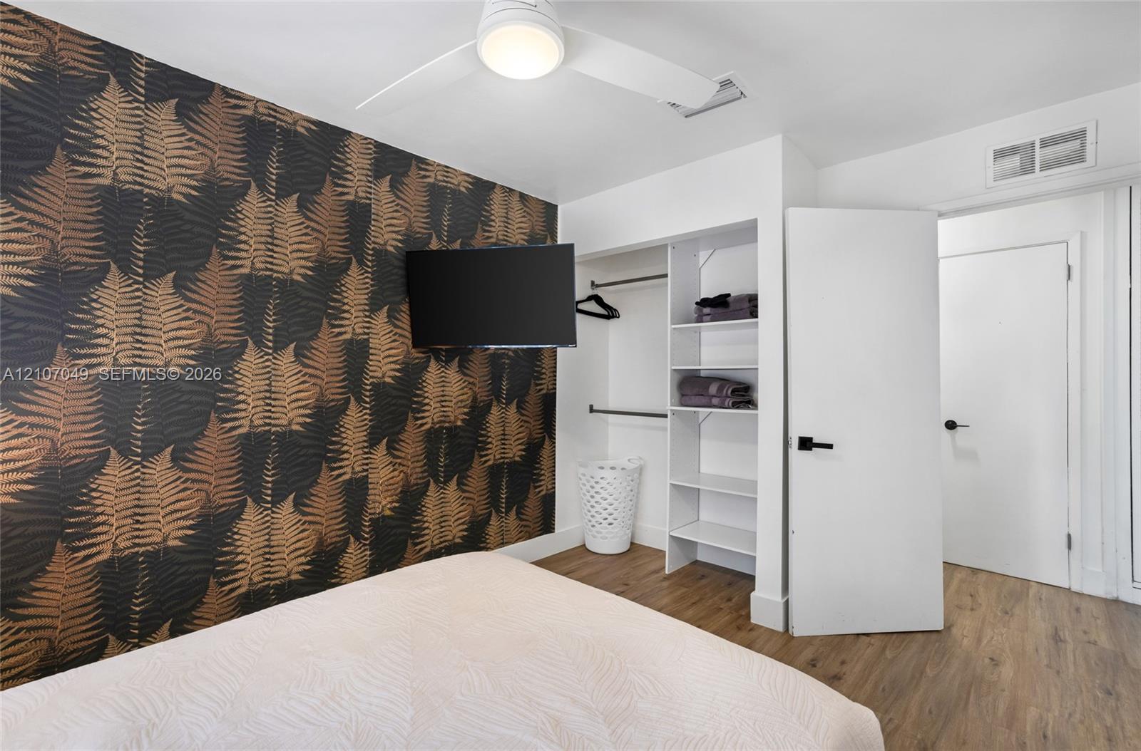 Accent wall with botanical wallpaper, wood-finish flooring, white ceiling fan with integrated lighting, and built-in closet featuring shelving and hanging rods