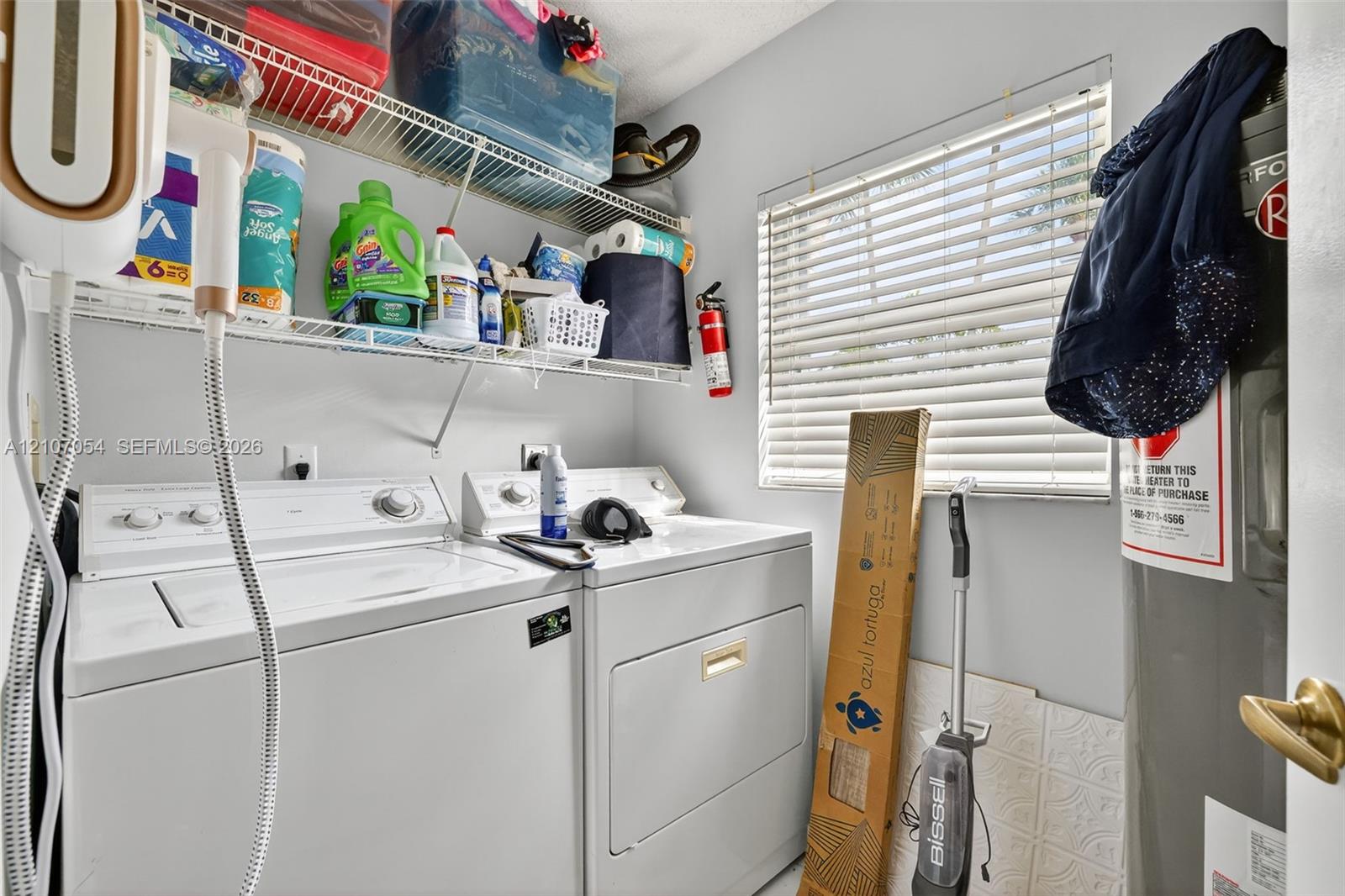 Laundry area featuring white top-load washer and dryer, horizontal window blinds, and tiered wire shelving