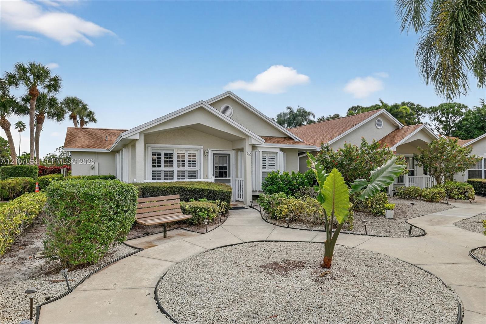 Single-story residence featuring a gable roof, light-toned exterior finish, shuttered windows, and concrete walkways through xeriscaped grounds