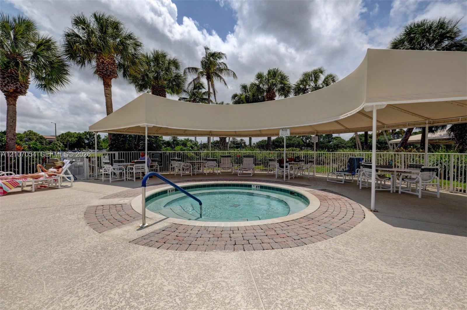 In-ground spa with a circular brick inlay, surrounded by a textured concrete deck, covered by a permanent curved canopy, and enclosed by white metal fencing