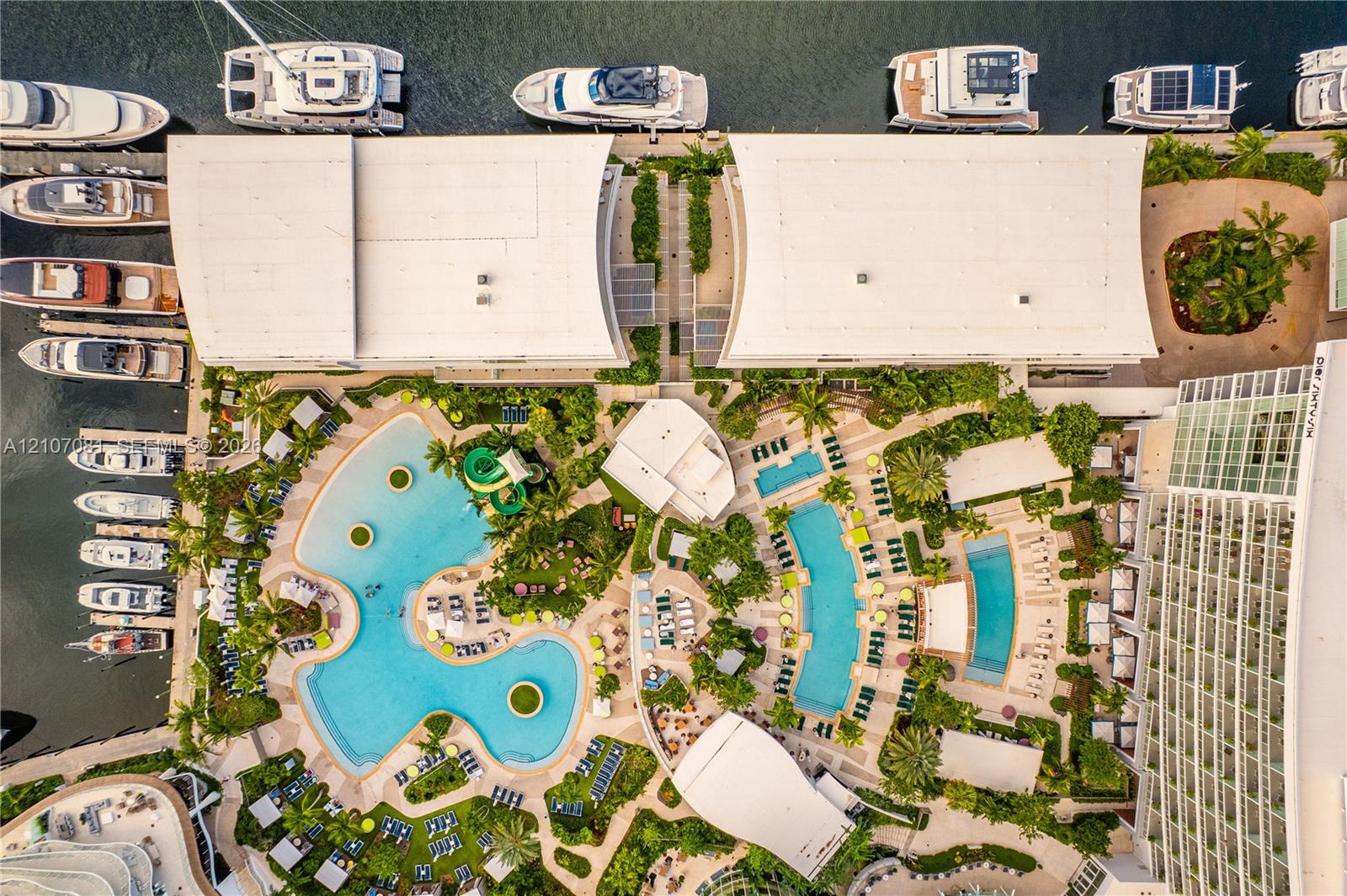 Aerial perspective of a waterfront property featuring multiple swimming pools, paved pool decks, lush landscaped grounds, and direct marina access