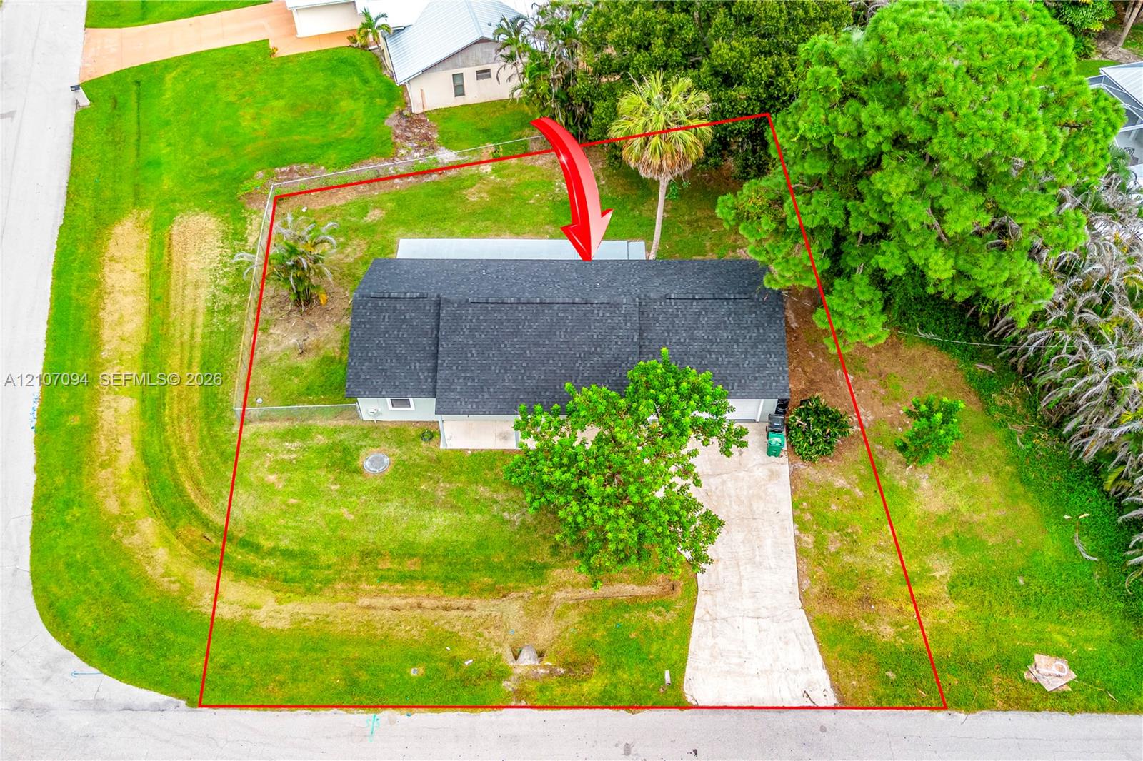 Aerial perspective of a property featuring a rectangular roofline, concrete driveway, fenced yard, and established landscaping including mature trees and palm specimens