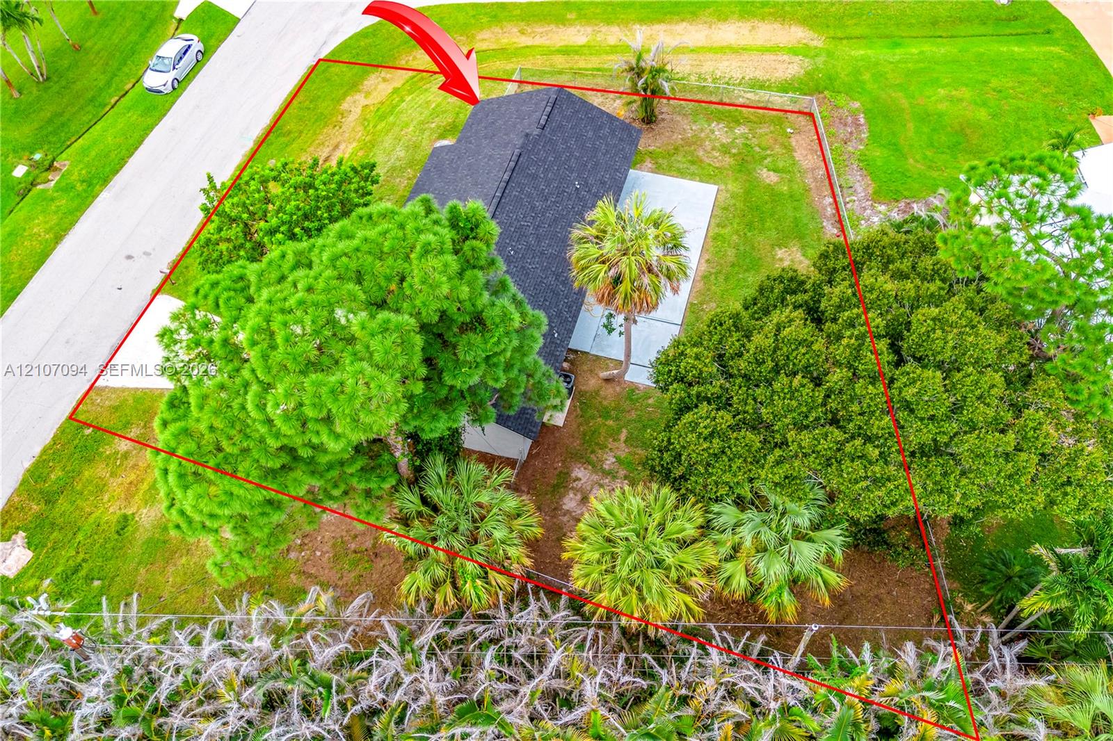 Aerial perspective of the property featuring a dark-toned shingled roof, a concrete patio, and a landscaped lot with mature palm and broadleaf trees