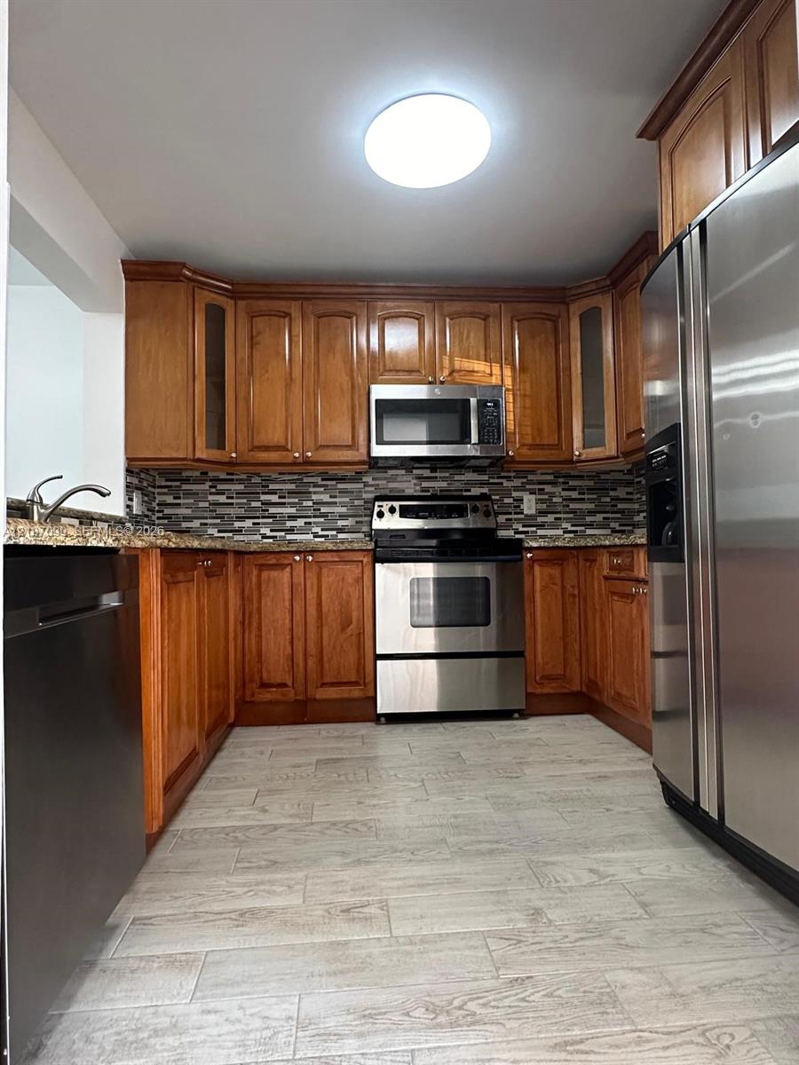 Kitchen featuring warm-toned wood cabinetry, multi-textured stone-look backsplash, stainless steel appliances, stone-finish countertops, and wood-finish flooring