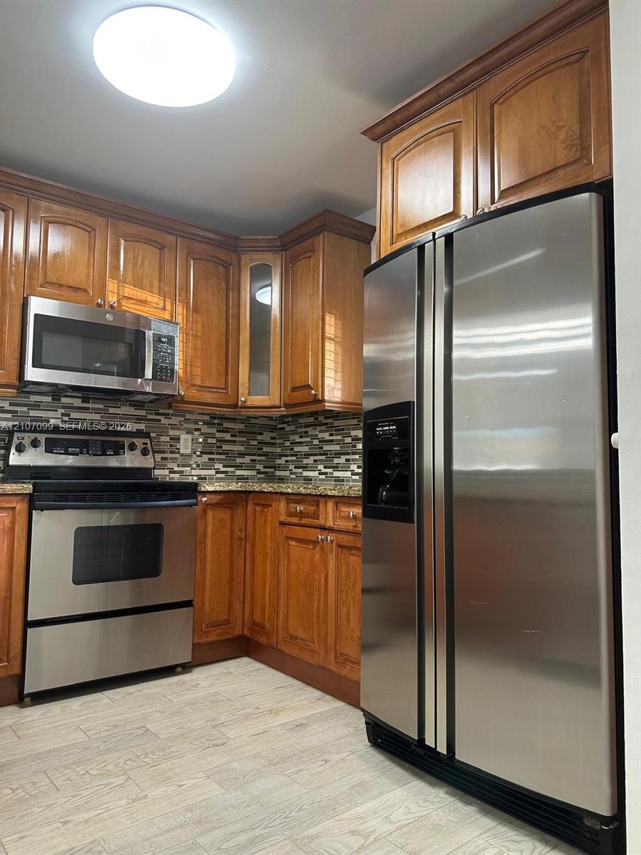 Kitchen featuring wood cabinetry, stainless steel appliances, stone-look countertops, a mosaic tile backsplash, and wood-finish flooring
