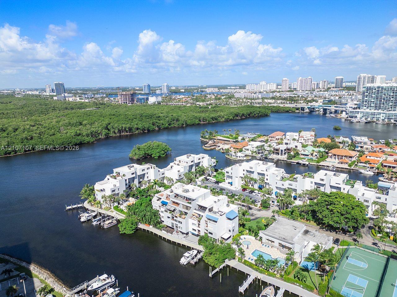 Aerial perspective of a waterfront residential complex featuring boat slips, a swimming pool, a tennis court, and dense coastal vegetation