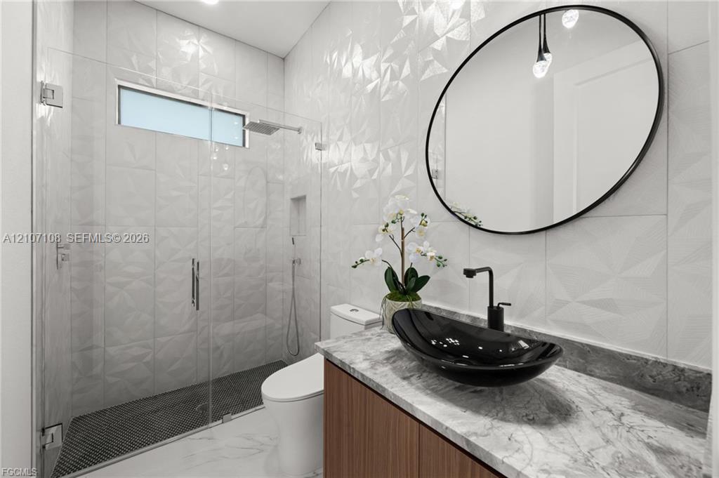 Bathroom featuring a custom wood-finish vanity with a stone-top surface, black vessel sink, geometric textured wall tile, frameless glass shower enclosure, and a circular black-framed mirror