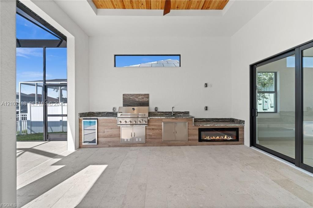 Covered lanai featuring a built-in outdoor kitchen with stainless steel appliances, stone-look countertops, wood-finish cabinetry, a linear fireplace, and tray ceiling with wood paneling