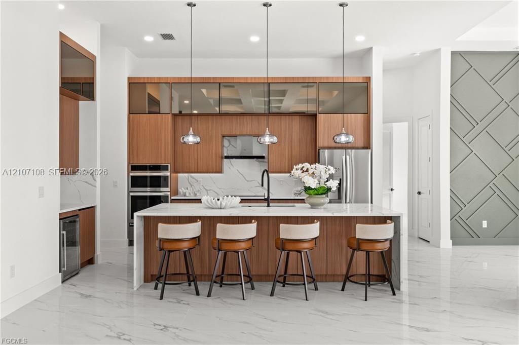 Modern kitchen featuring flat-panel wood cabinetry, a central island with a waterfall countertop, stone-finish backsplash, three glass-pendant lights, and geometric wall molding Modern kitchen featuring flat-panel wood cabinetry, a central island with a waterfall countertop, stone-finish backsplash, three glass-pendant lights, and geometric wall molding