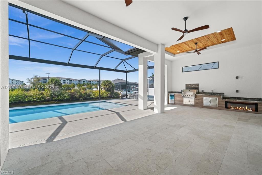 Covered outdoor kitchen featuring built-in stainless steel appliances, linear fireplace, wood-plank ceiling accent, and stone-tile flooring opening to a screened-in swimming pool