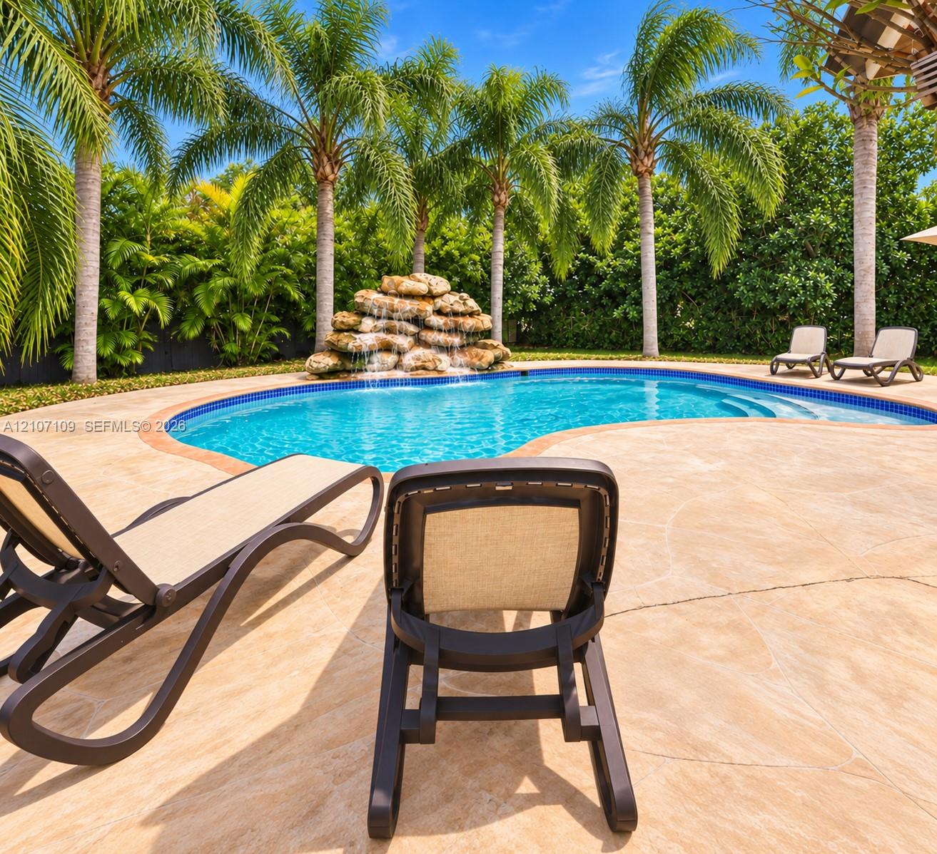 Free-form swimming pool featuring a stacked-stone waterfall, integrated steps, and stone-finish patio surfacing, surrounded by mature palm trees