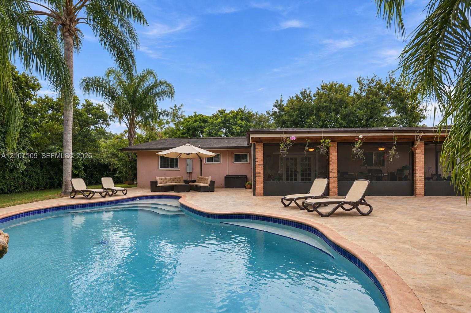 Outdoor living space featuring a free-form pool, expansive paved patio, covered screened-in area, brick structural pillars, and mature palm trees