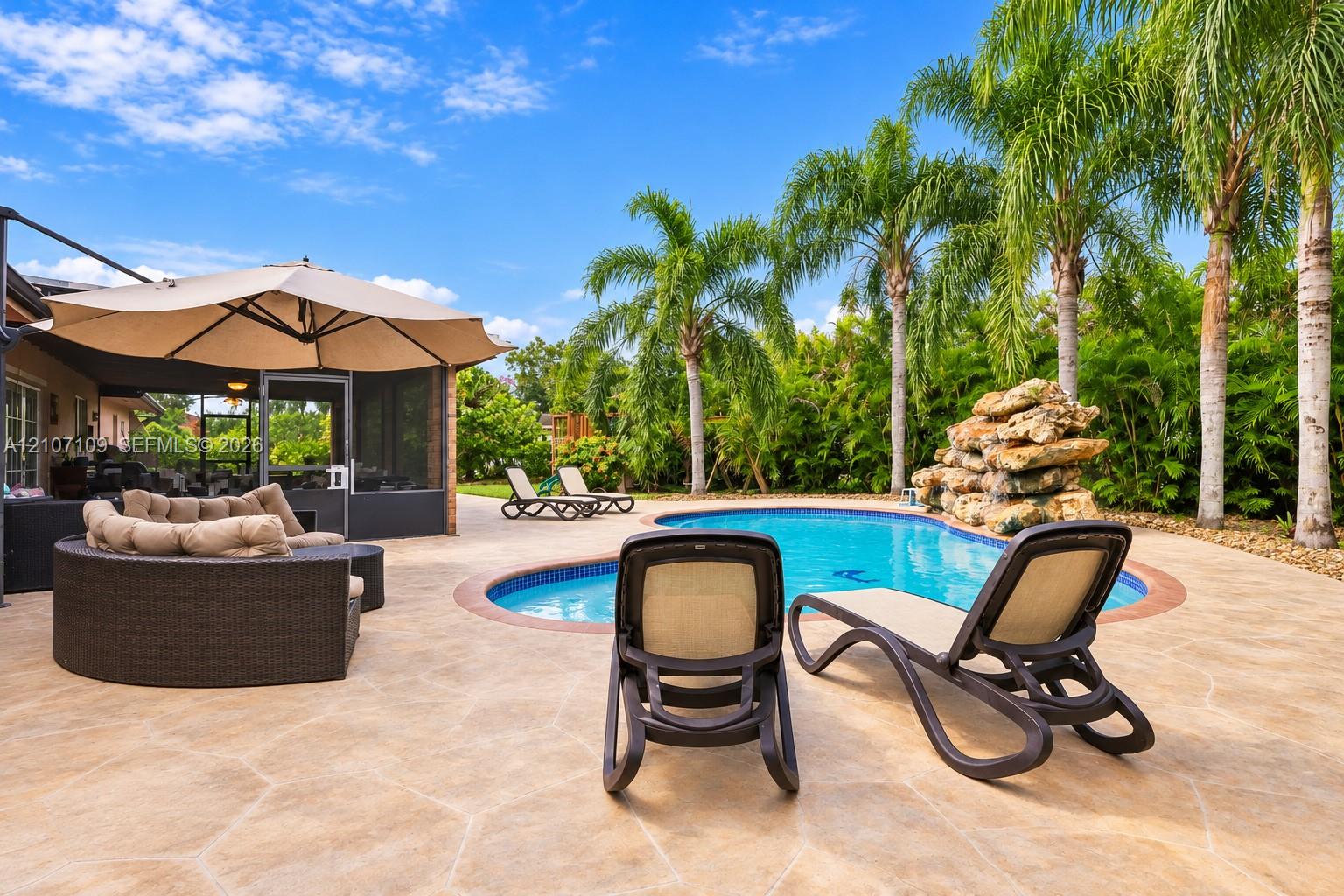 Outdoor patio featuring stamped concrete hardscaping, a swimming pool with tile trim, a stone waterfall feature, and established palm trees