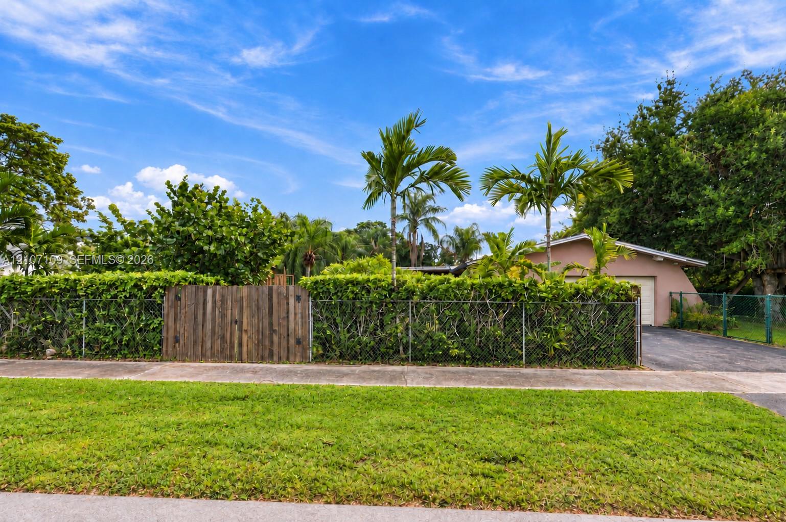 Exterior grounds featuring a metal chain-link perimeter fence, a solid wood privacy gate, mature tropical landscaping, and an asphalt driveway