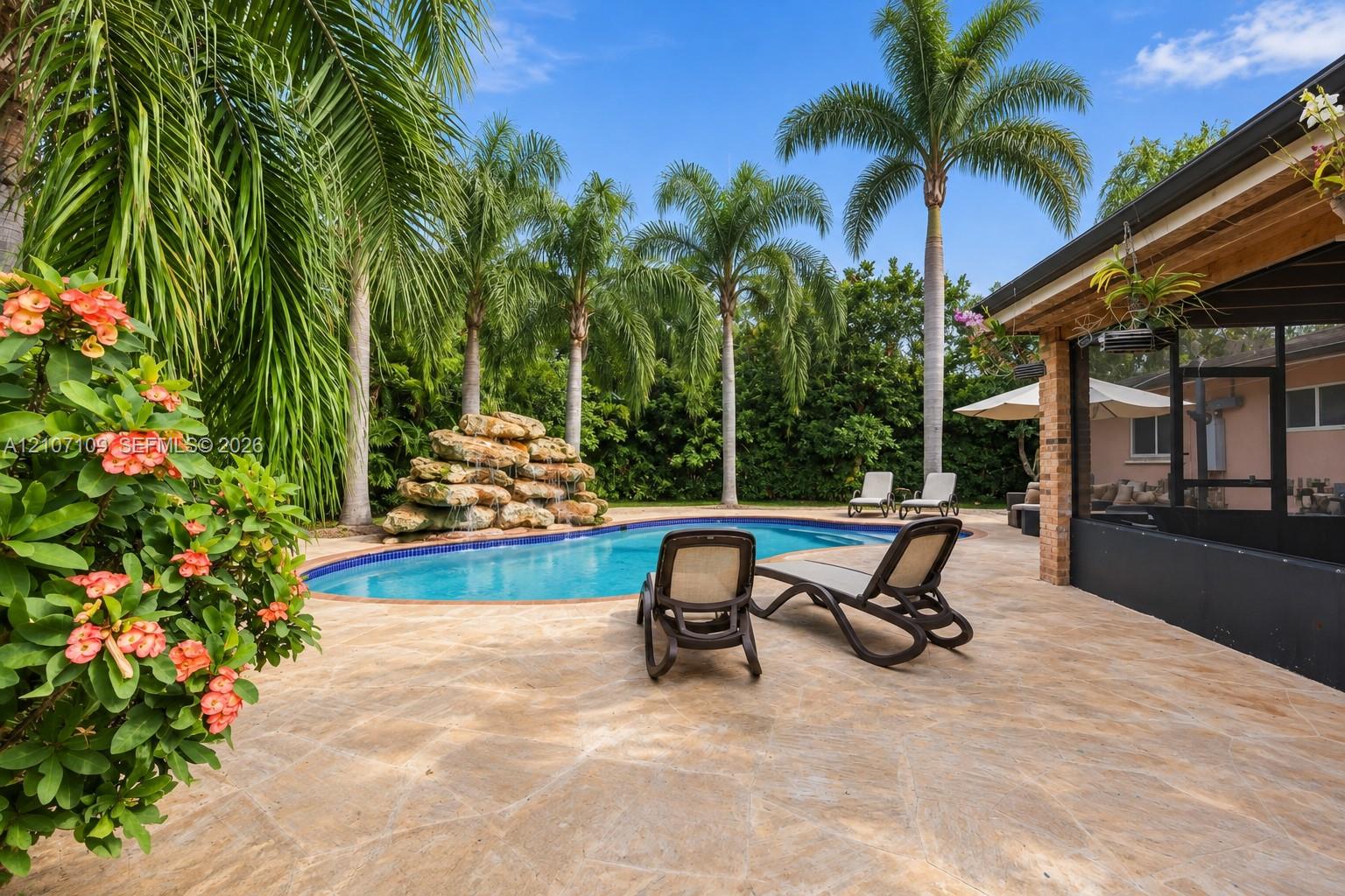In-ground swimming pool featuring a stacked stone water feature, stone-paver decking, and mature palm trees