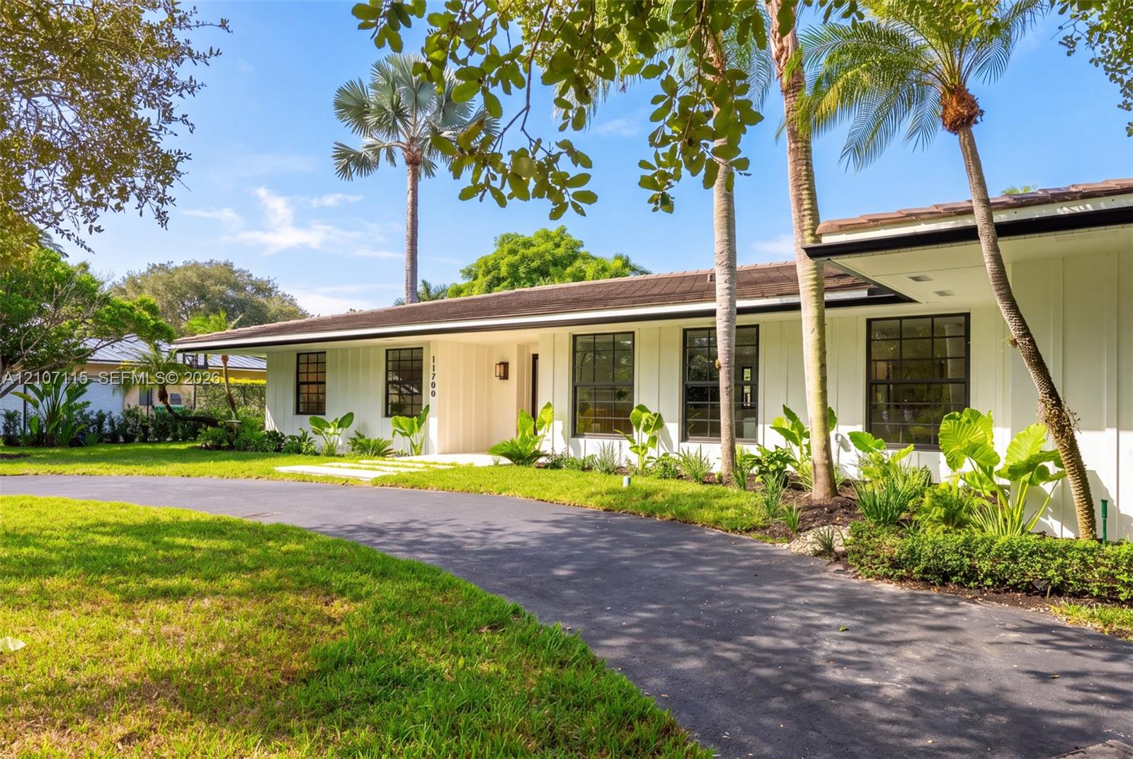 Single-story exterior featuring board-and-batten siding, a low-pitched roof, multi-pane windows, and a curved asphalt driveway surrounded by mature palm trees and manicured landscaping