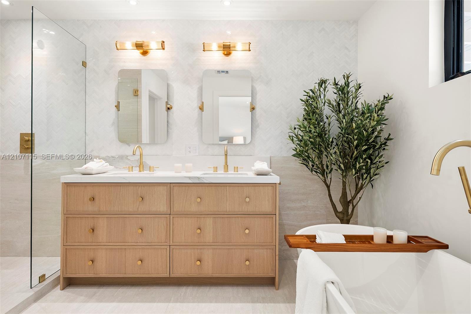 Double vanity featuring light wood cabinetry with gold-toned hardware, stone countertops, dual rectangular mirrors, and a glass-enclosed shower with herringbone wall tiling
