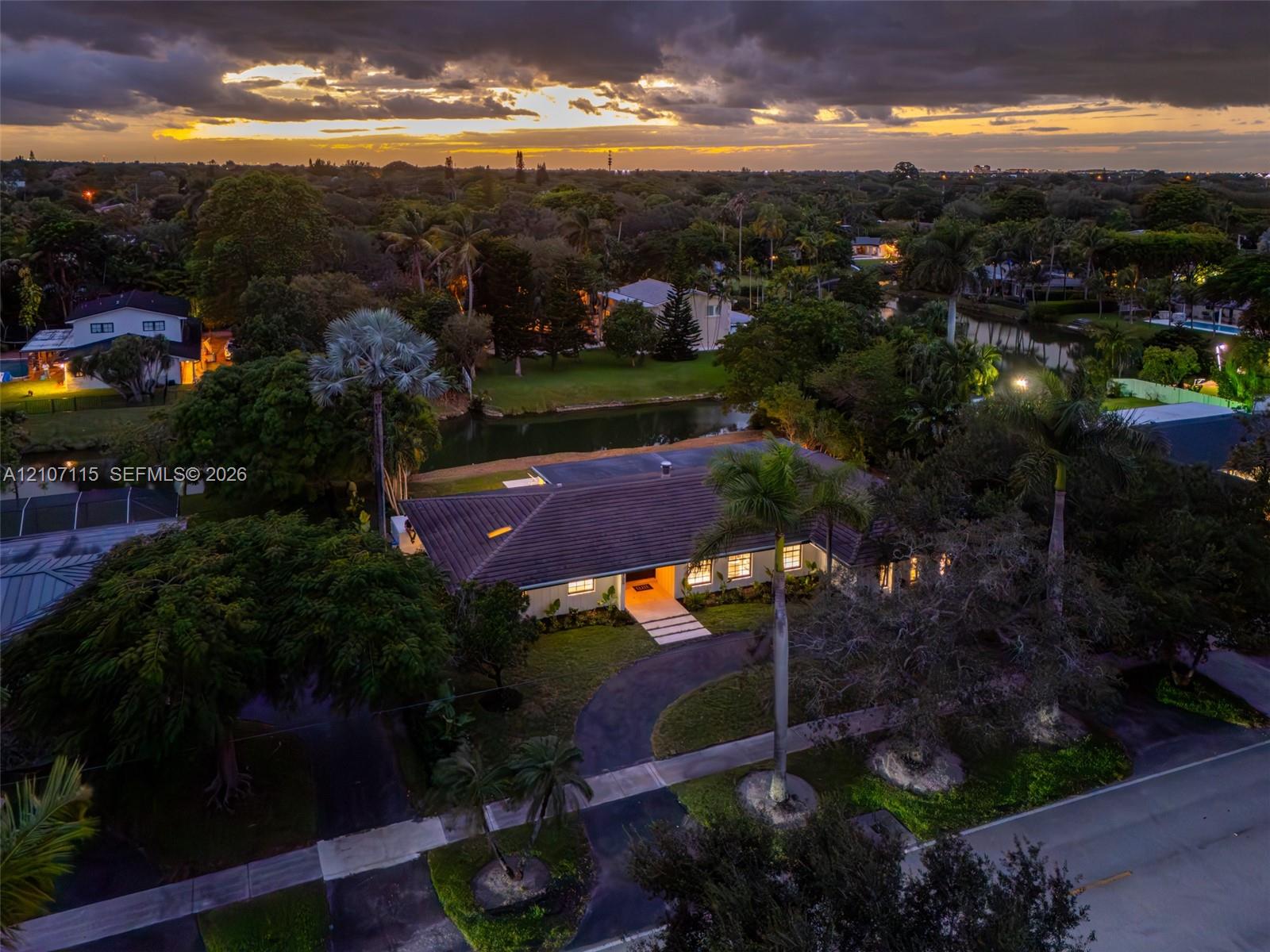 Aerial perspective featuring a low-slope roof structure, a paved circular driveway, manicured landscaping with mature palm trees, and direct waterfront access