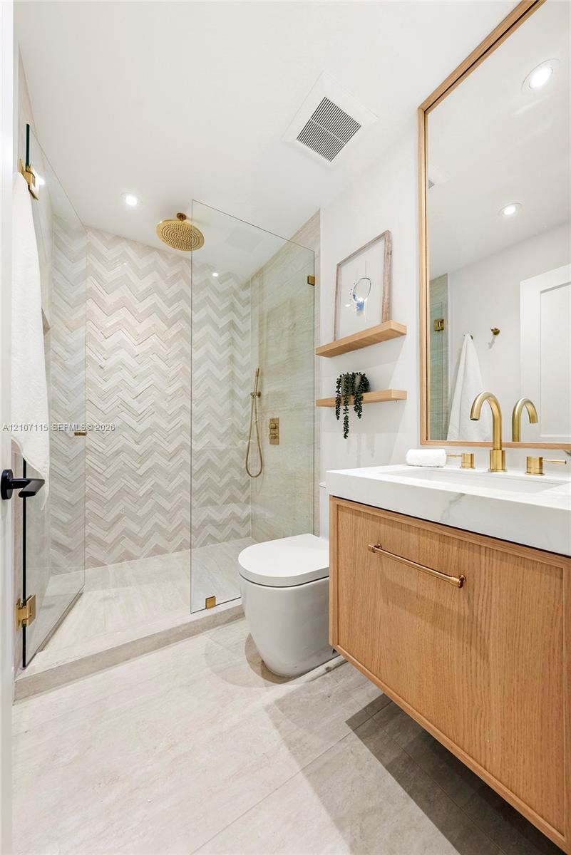 Wood-finish floating vanity with a white countertop and brass hardware, glass-enclosed walk-in shower with herringbone tile walls, and a large wall-mounted mirror with a wood frame