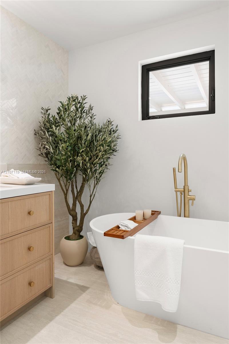 Freestanding soaking tub with a brass floor-mounted faucet, light wood-finish cabinetry, white countertop, herringbone-patterned wall tile, and a black-framed window