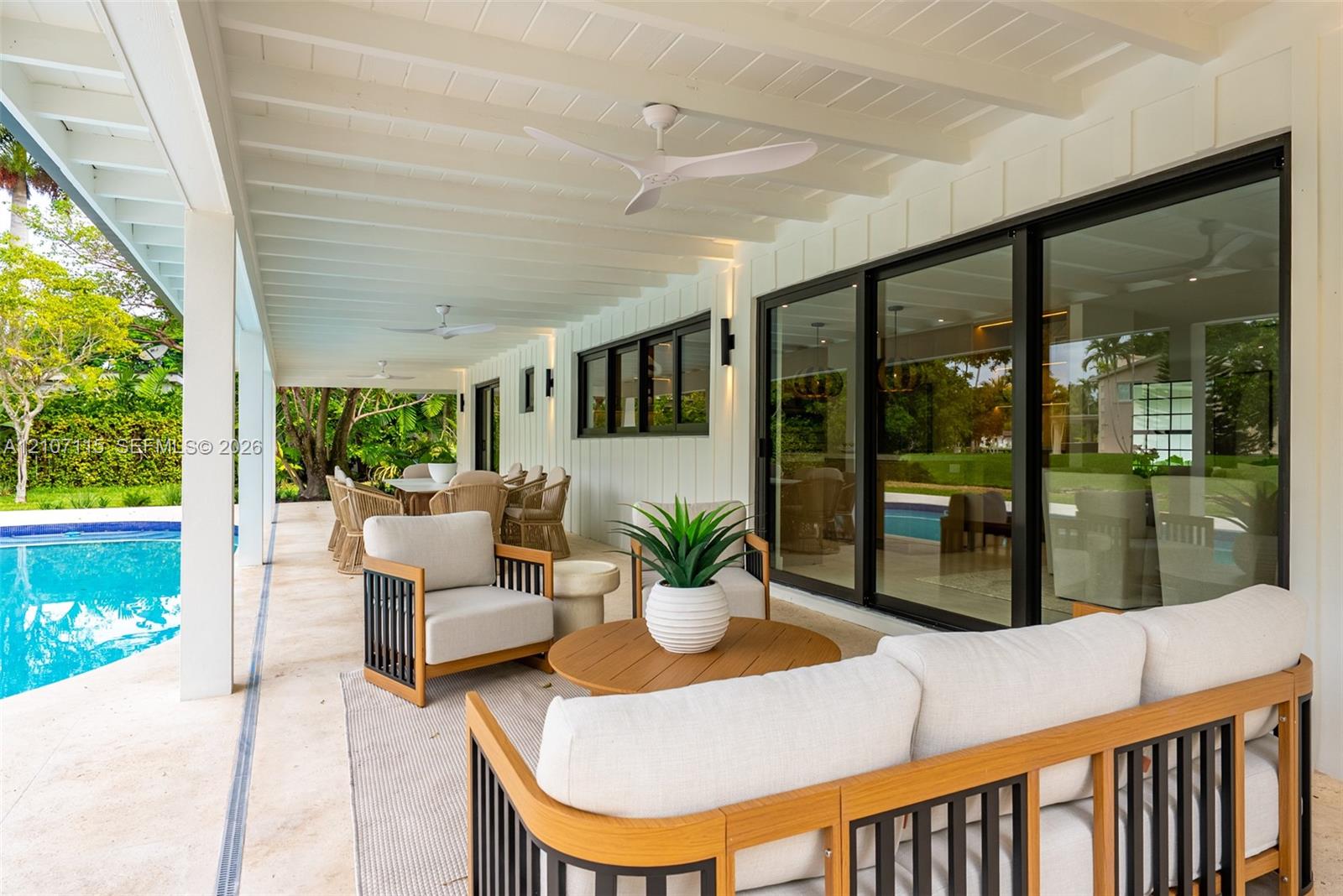 Covered outdoor patio featuring white tongue-and-groove ceiling, dual modern ceiling fans, sliding glass wall panels, and stone-finish flooring overlooking a private swimming pool