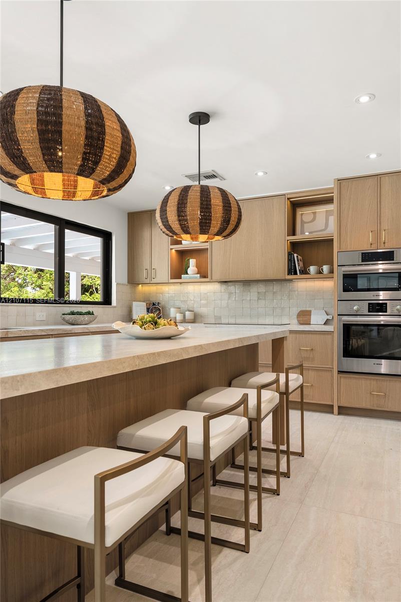 Custom wood-finish cabinetry, stone-topped kitchen island, built-in wall oven and microwave, textured tile backsplash, and oversized woven pendant lighting