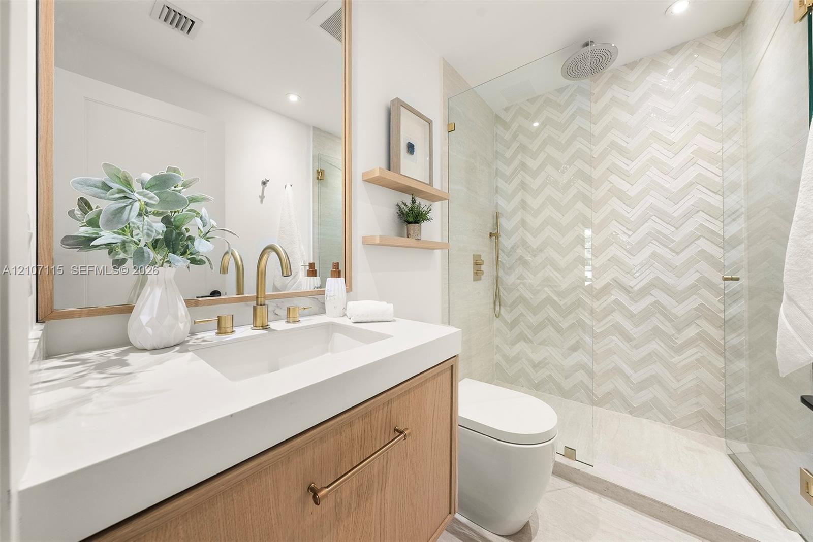 Bathroom featuring a wood-finish vanity with a quartz countertop, brass-toned fixtures, a walk-in shower with chevron-patterned tile, and two floating wooden shelves