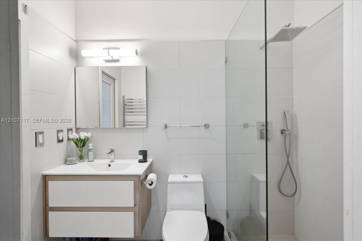Floating wood-finish vanity with a white countertop, walk-in shower with a glass panel, rainfall shower head, handheld sprayer, and wall-mounted vanity lighting