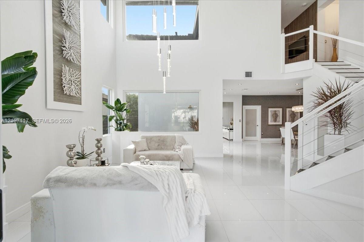 Double-height ceiling, large-format polished tile flooring, staircase with cable railing, tiered contemporary chandelier, and expansive picture windows