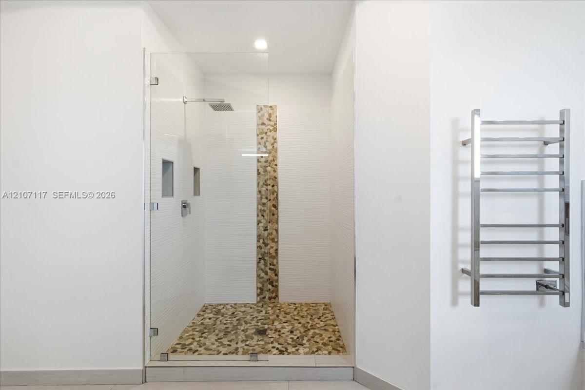 Walk-in shower featuring a frameless glass door, stone-tile flooring and wall accent, dual recessed wall niches, rainfall showerhead, and a wall-mounted metal towel warmer