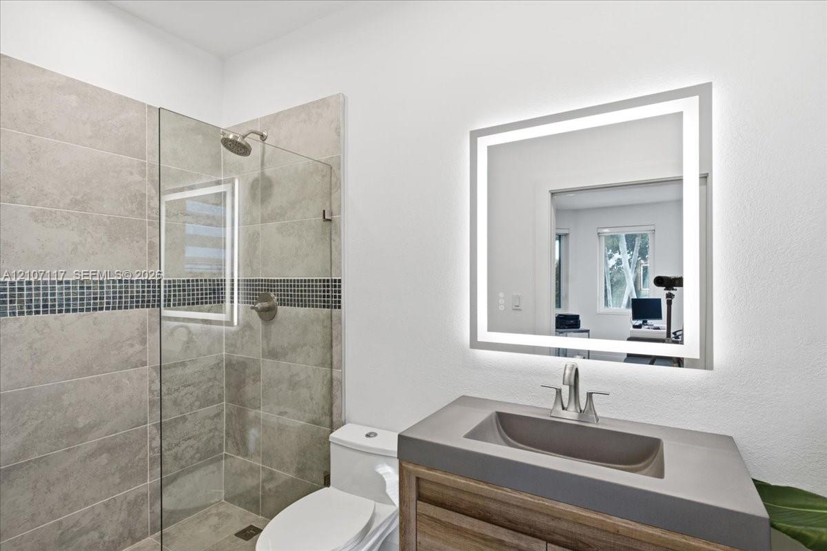 Walk-in shower with stone-finish tile and a mosaic tile inlay, wood-finish vanity with a molded sink, and a wall-mounted LED-backlit mirror