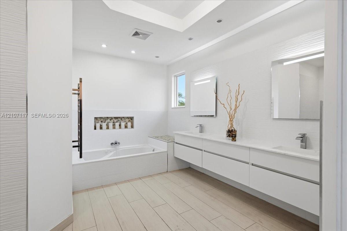 Modern bathroom featuring a long floating dual-sink vanity with a solid-surface countertop, an inset tiled soaking tub with a decorative stone-accented recessed niche, wood-finish flooring, and wall-mounted mirrors with integrated lighting