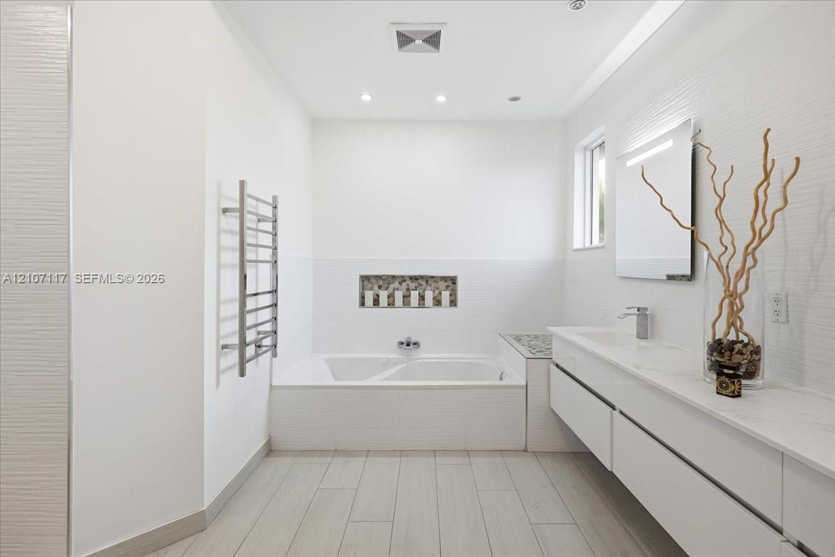 Floating double vanity with quartz countertop, inset soaking tub featuring a decorative stone mosaic niche, wood-finish tile flooring, and wall-mounted heated towel rack